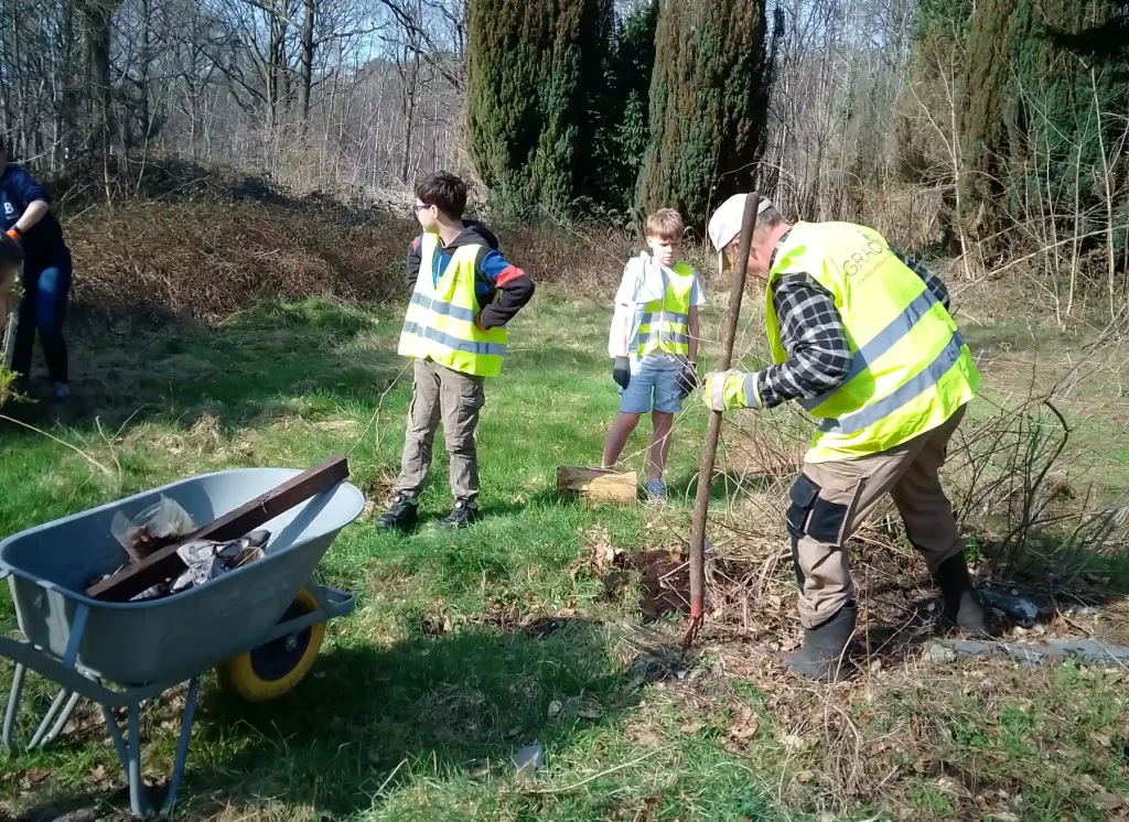 Groupe de personnes portant des gilets réfléchissants travaillant à l’extérieur dans un espace vert. Une personne utilise un outil pour nettoyer ou dégager des branches au sol, tandis que deux autres observent et participent à l’activité. Une brouette remplie de matériaux se trouve au premier plan