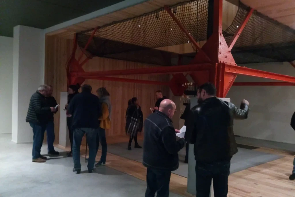Groupe de personnes discutant dans une salle d’exposition autour d’une grande structure métallique rouge avec filet suspendu, installée sur un sol en bois clair.