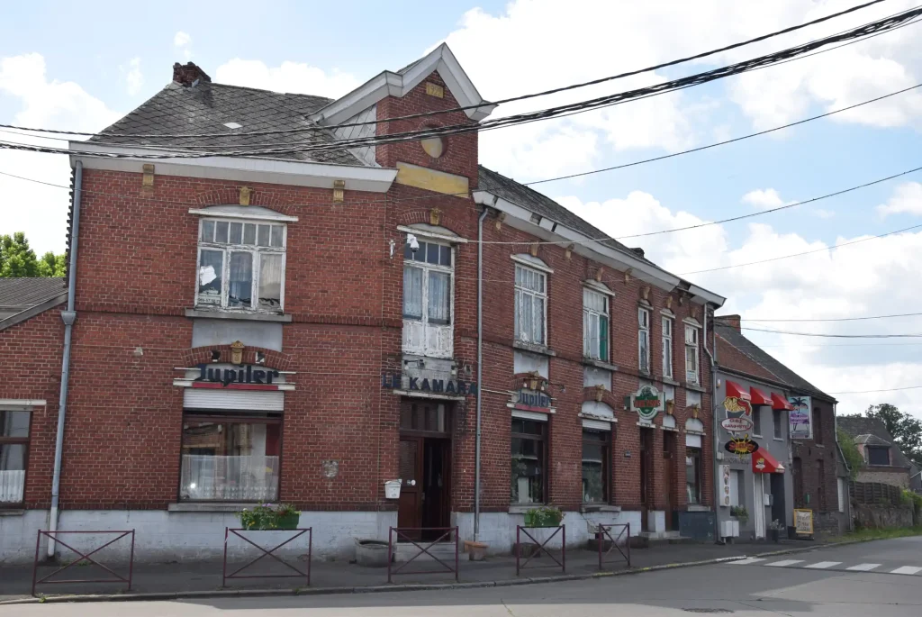 Bâtiment en briques rouges à deux étages avec vitrines et enseignes commerciales, situé à l’angle d’une rue, avec une voiture blanche garée au premier plan.