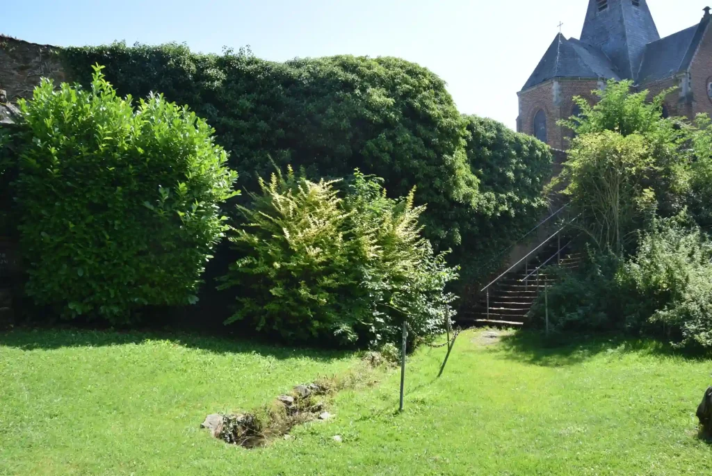 Jardin verdoyant avec pelouse, arbustes et haies denses, au pied d’un escalier en pierre menant à une église en briques avec toit en ardoise