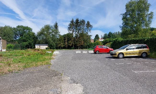 Petit parking extérieur ensoleillé, bordé d’arbres et de haies, avec deux voitures stationnées sur la droite.
