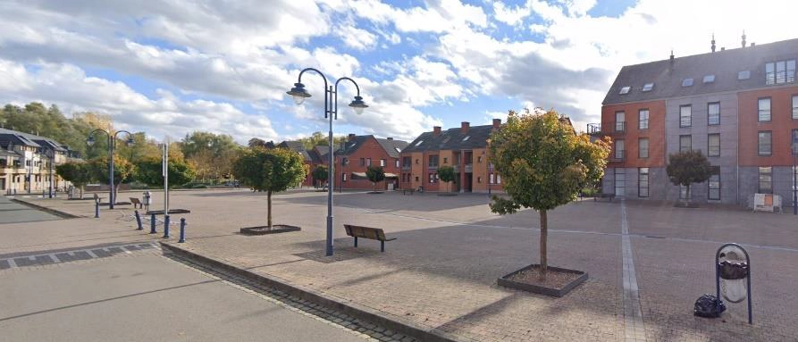 Grande place urbaine pavée, bordée d’arbres et de bâtiments en briques, avec bancs et lampadaires sous un ciel nuageux.