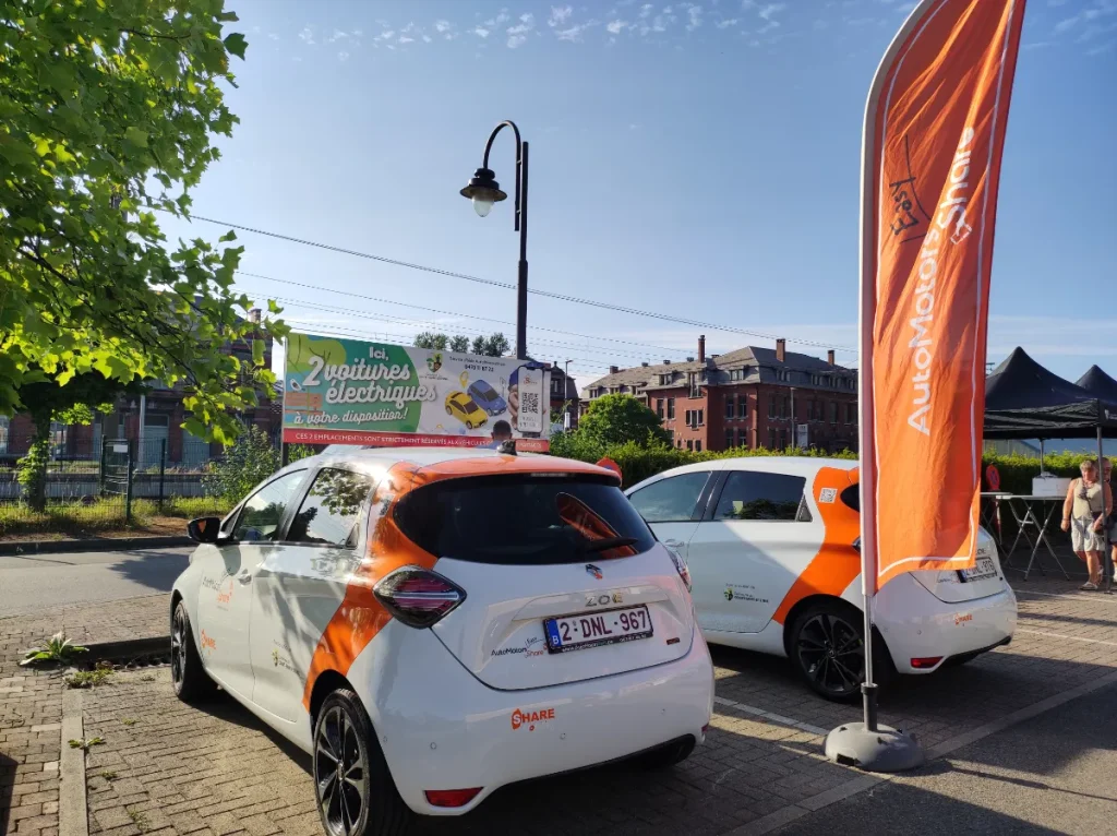 Deux voitures électriques en libre‑partage stationnées sur une place, à côté d’un drapeau promotionnel et d’un panneau annonçant le service.