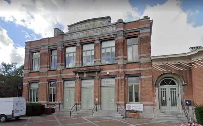 Façade en briques du bâtiment du Foyer Populaire, avec grandes portes vert clair, escaliers d’accès et enseignes associatives.