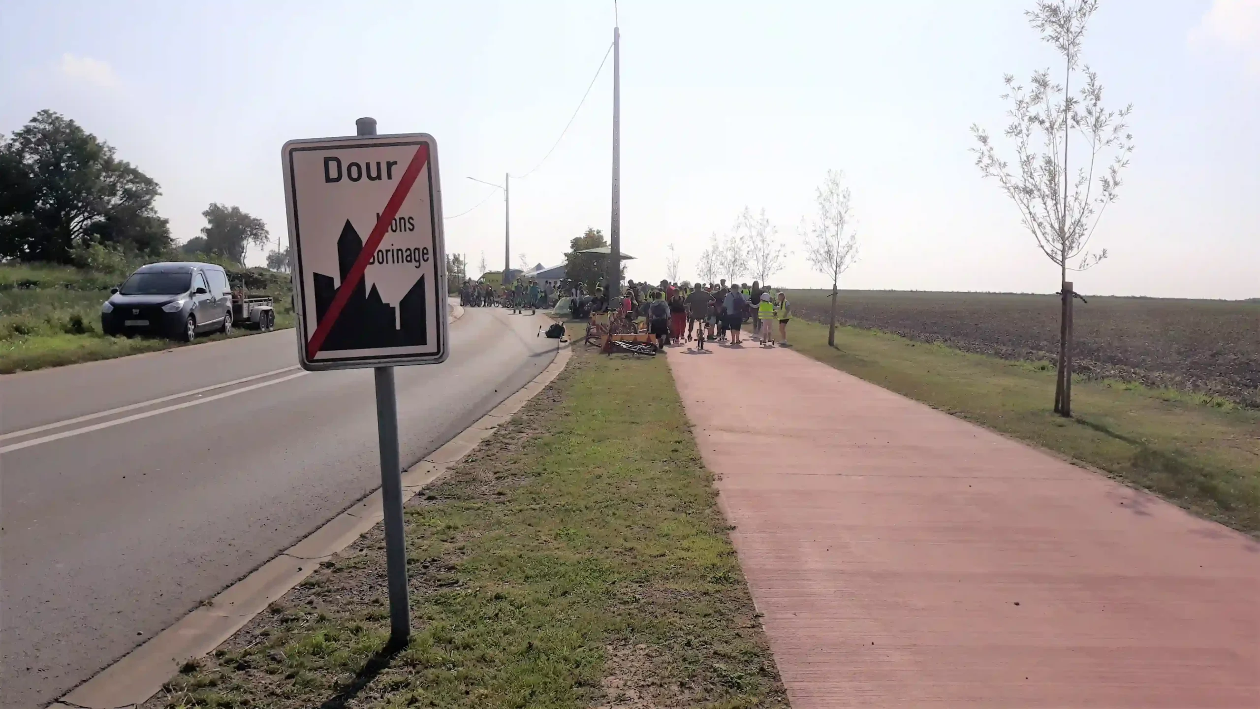 Panneau indiquant la fin de l’agglomération de Dour le long d’une route, avec une piste cyclable et un groupe de personnes rassemblées à l’arrière-plan.