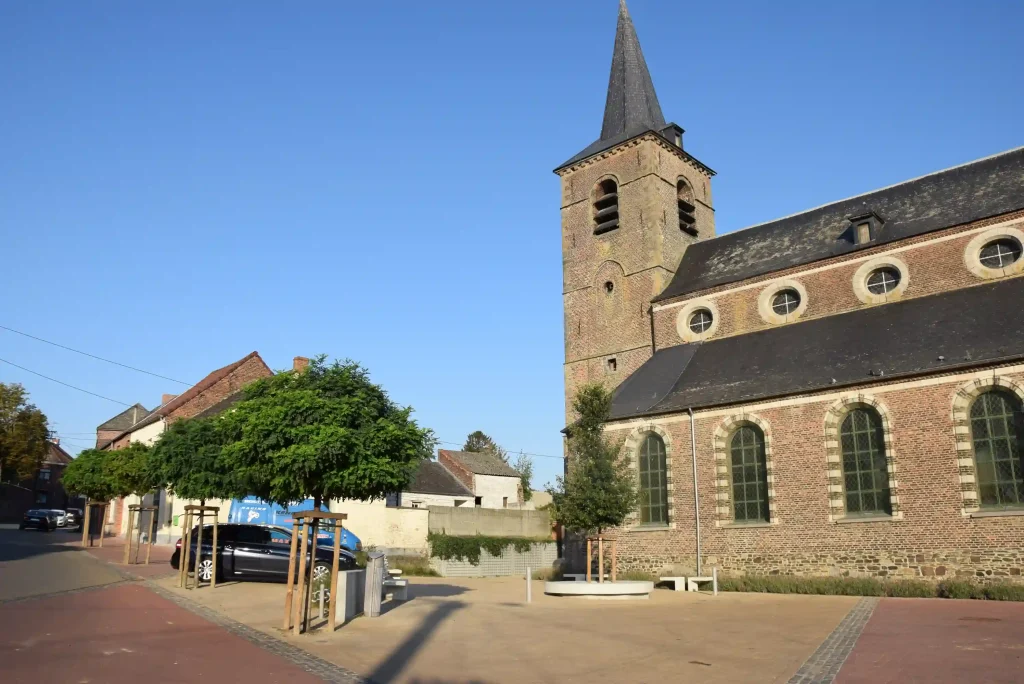 Église en briques avec clocher et place aménagée avec jeunes arbres.