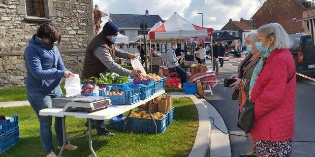 Marché de plein air avec des stands de fruits et légumes. Des vendeurs disposent des produits dans des caisses tandis que des personnes portant des masques attendent devant la table pour acheter, dans un village avec des bâtiments en pierre en arrière-plan