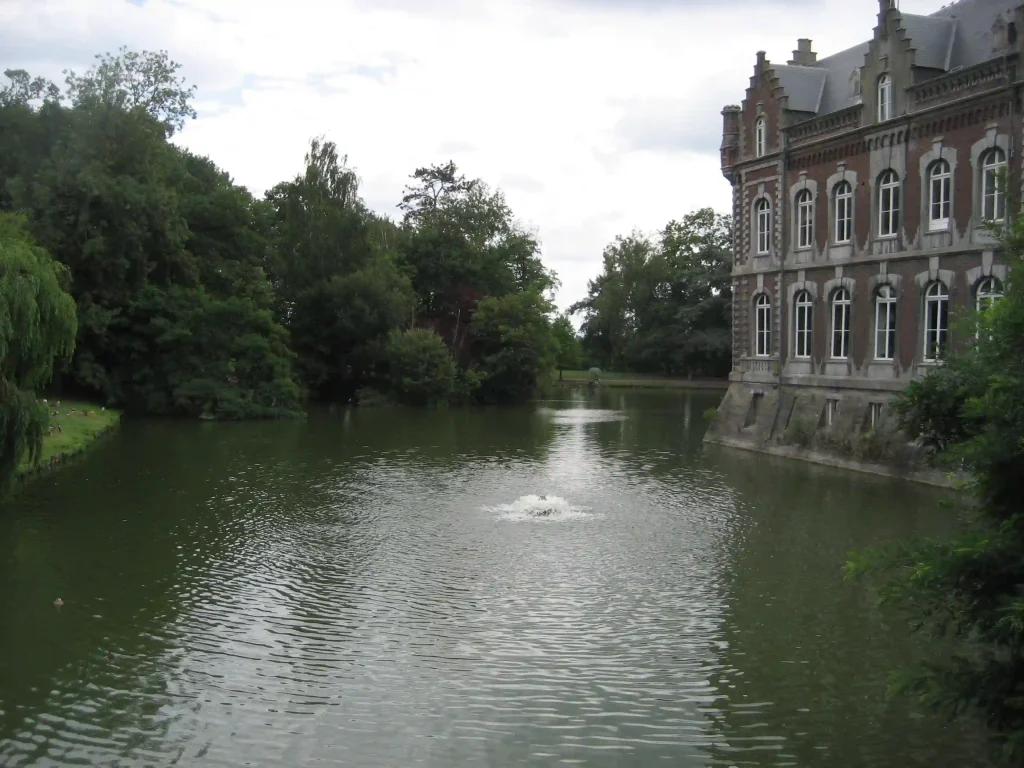 Château en briques avec tourelles, bordé d’un étang arboré avec fontaine.