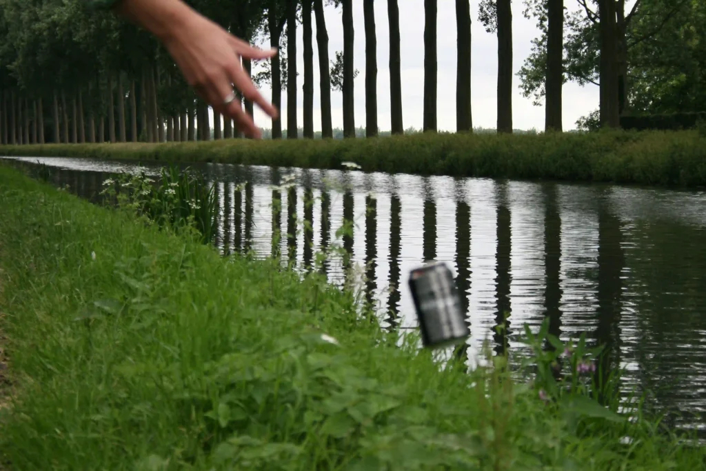 Main lançant une canette dans un canal bordé d’arbres, geste de pollution de l’environnement.