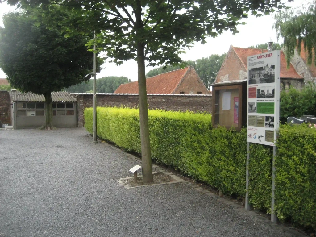 Allée gravillonnée bordée de haies avec panneau d’information sur Saint‑Léger et bâtiments en briques à l’arrière.