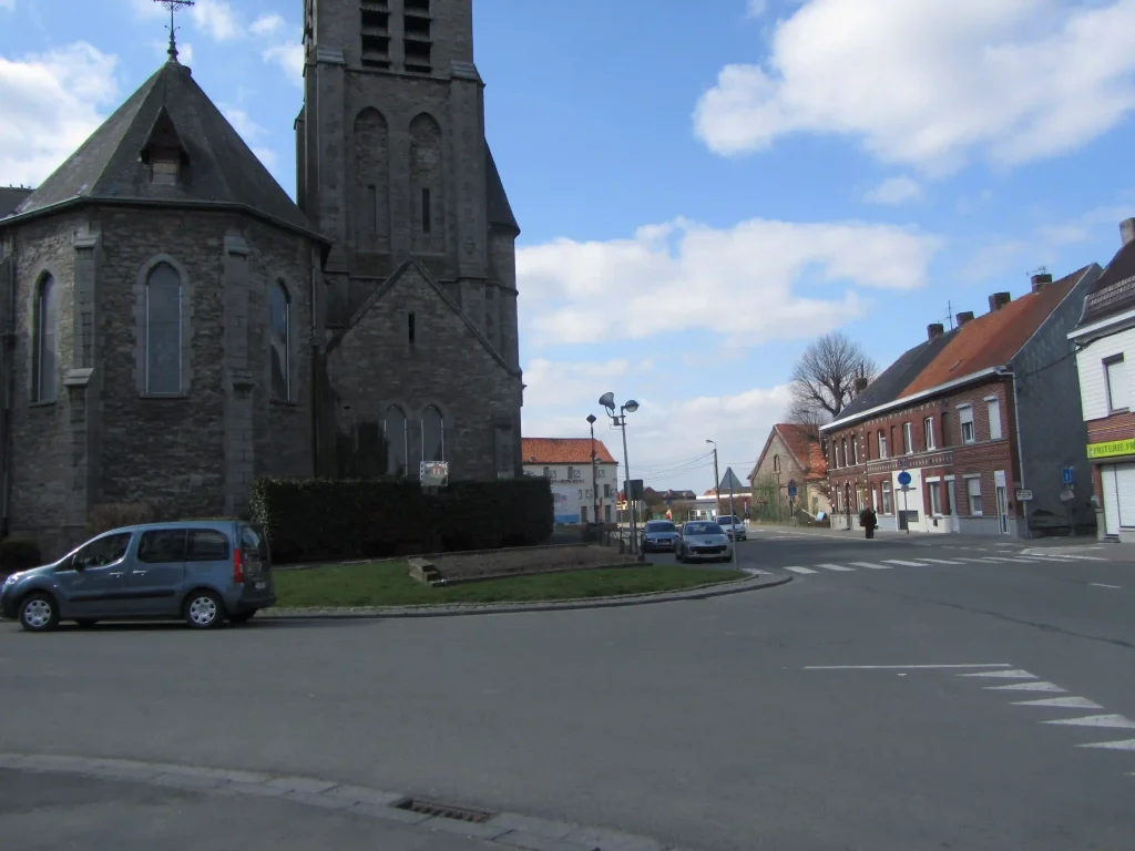 Église en pierre et carrefour de village avec maisons en briques et voitures stationnées.