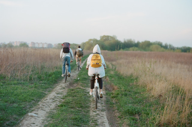 Trois personnes à vélo circulent sur un chemin de terre au milieu d’un paysage herbeux, avec des sacs à dos, sous un ciel dégagé.
