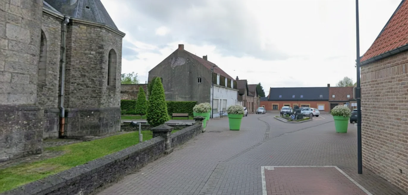 Place de village pavée avec bâtiment en pierre à gauche, maisons en briques, quelques voitures et grands pots de fleurs verts sous un ciel nuageux.