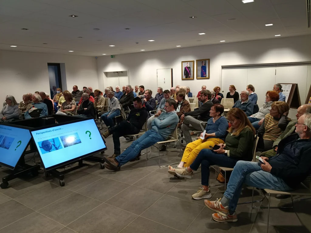 Réunion publique dans une salle : public assis face à un écran de présentation.
