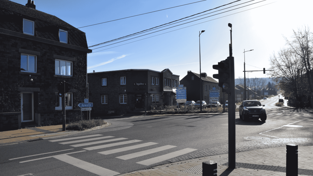 Carrefour routier réaménagé à Manhay avec passages pour piétons, feux de signalisation et panneaux directionnels, bordé de bâtiments en pierre.