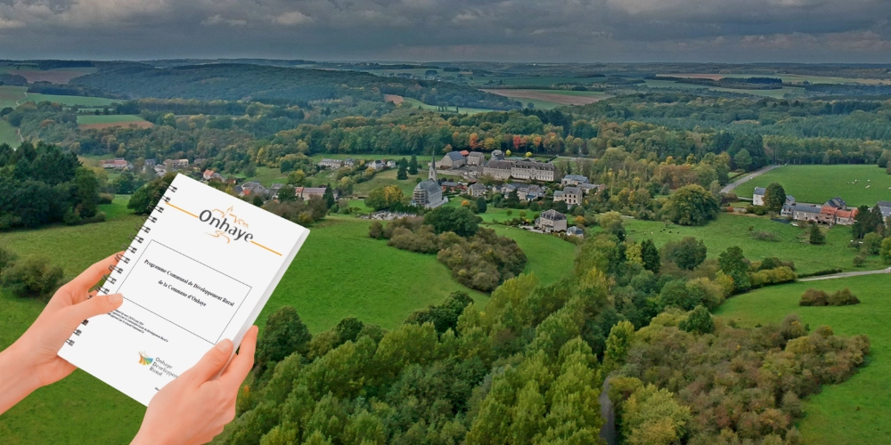 deux mains tiennent le pcdr de Onhaye en avant plan et une vue de Onhaye en arrière-plan