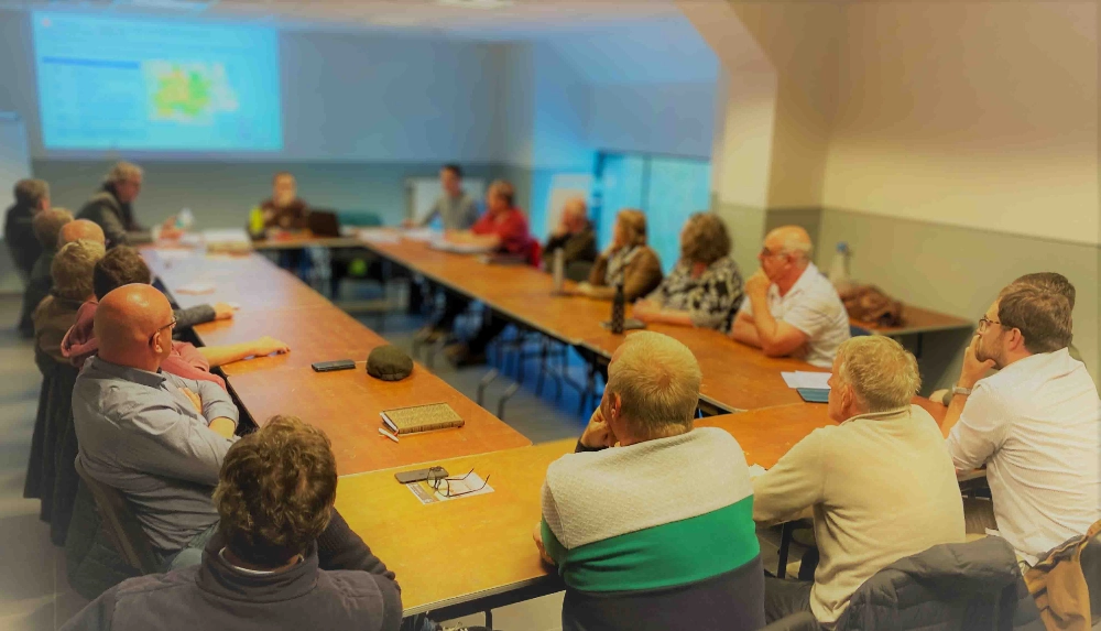 Une vingtaine de personne assise autour de tables disposées en un grand rectangle qui regardent une présentation projetée sur un mur en arrière plan de l'image