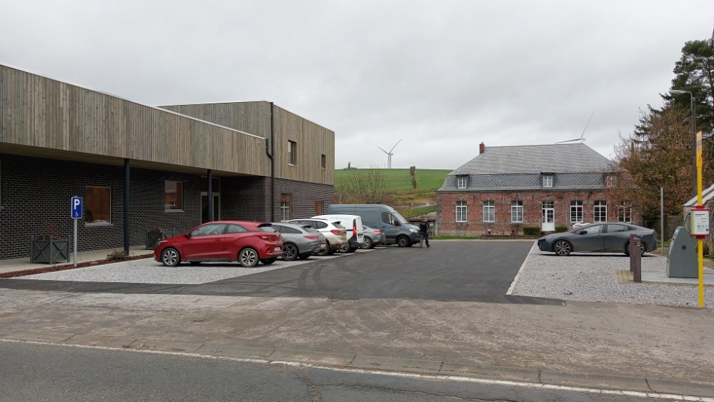 Nouvelle Maison de Village de Grandrieu avec façade en bois et briques, parking aménagé et bâtiment ancien en arrière-plan sous ciel gris.