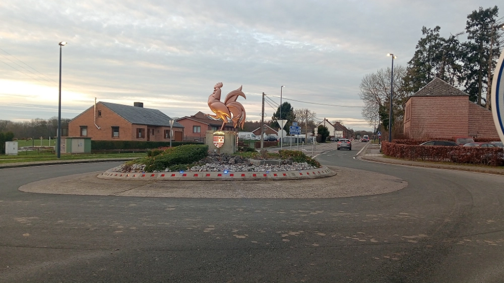 Vue du rond-point de Sivry-Rance après l’installation d’une infrastructure de franchissement. Au centre, une sculpture de coq trône sur un massif de pierres, entourée de routes et de maisons en briques.