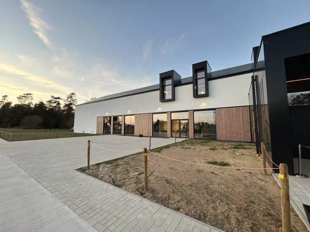 Façade moderne de la maison rurale Tintigny-Bellefontaine. Un bâtiment blanc et noir avec grandes baies vitrées, vue en fin de journée. Une esplanade pavée se trouve au premier plan