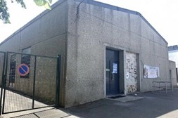 Bâtiment en béton gris avec une entrée centrale, entouré d’une clôture métallique, vu depuis l’extérieur.