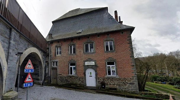 Bâtiment ancien en briques et pierres adossé à un pont en arc, avec une porte centrale et plusieurs fenêtres, situé le long d’une route et entouré d’arbres