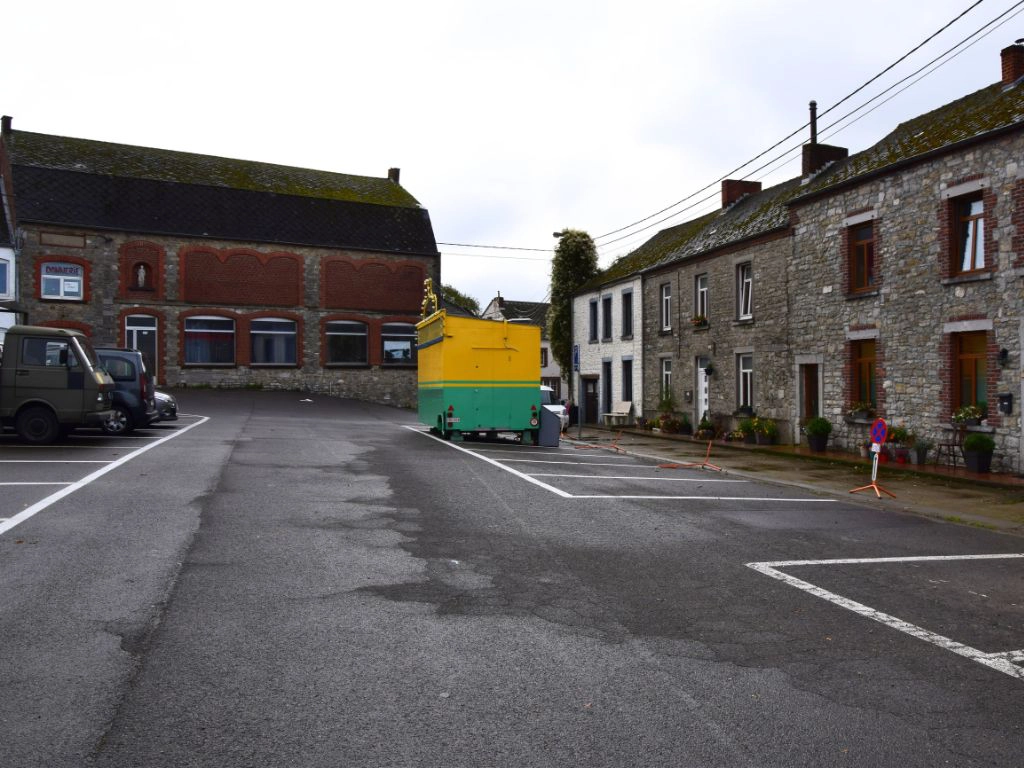 Vue de la place Saint-Walhère à Gourdinne (Walcourt), avec des maisons en pierre, un bâtiment communal et un équipement temporaire installé sur le parking