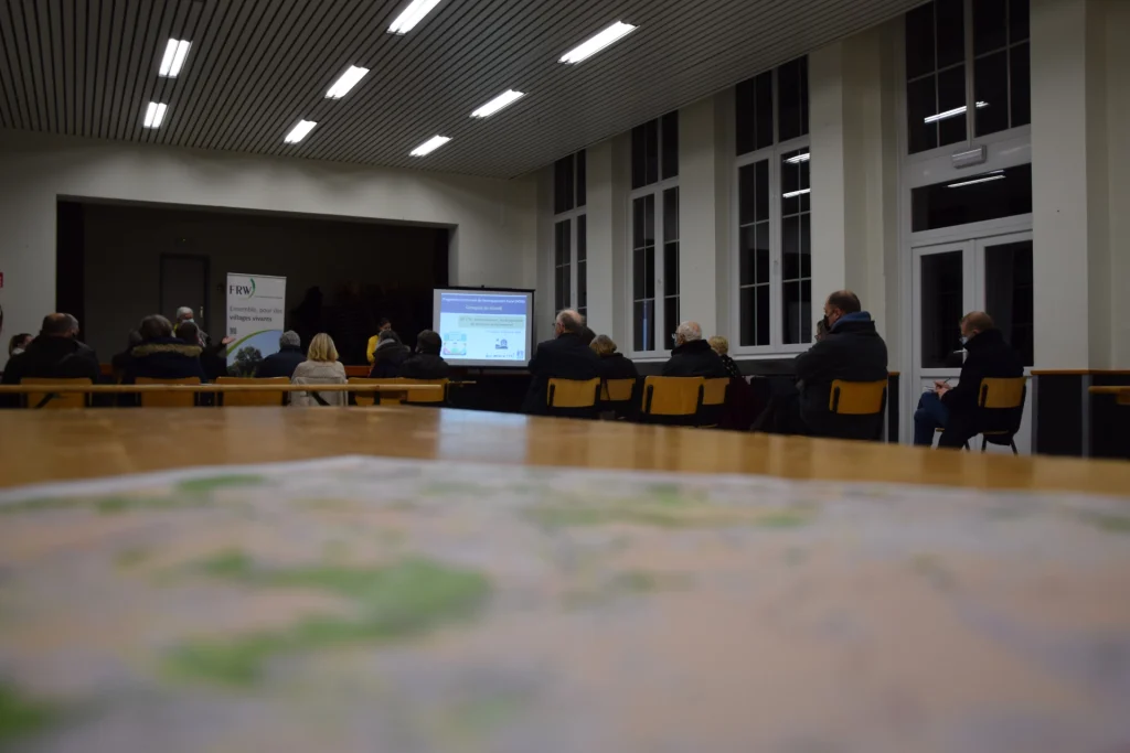 Réunion publique dans une salle avec des participants assis face à un écran de projection, vue depuis une table floue au premier plan.
