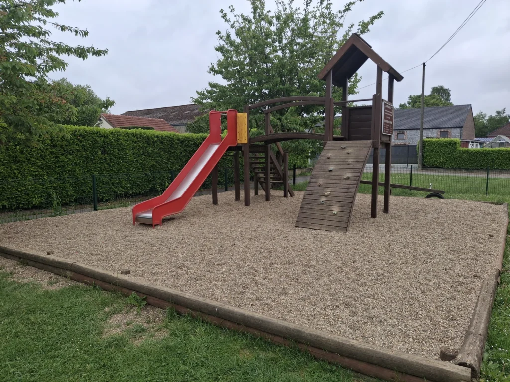 Aire de jeux extérieure avec un module en bois comprenant un toboggan rouge, une passerelle et un mur d’escalade pour enfants, installée sur un sol amortissant en graviers.