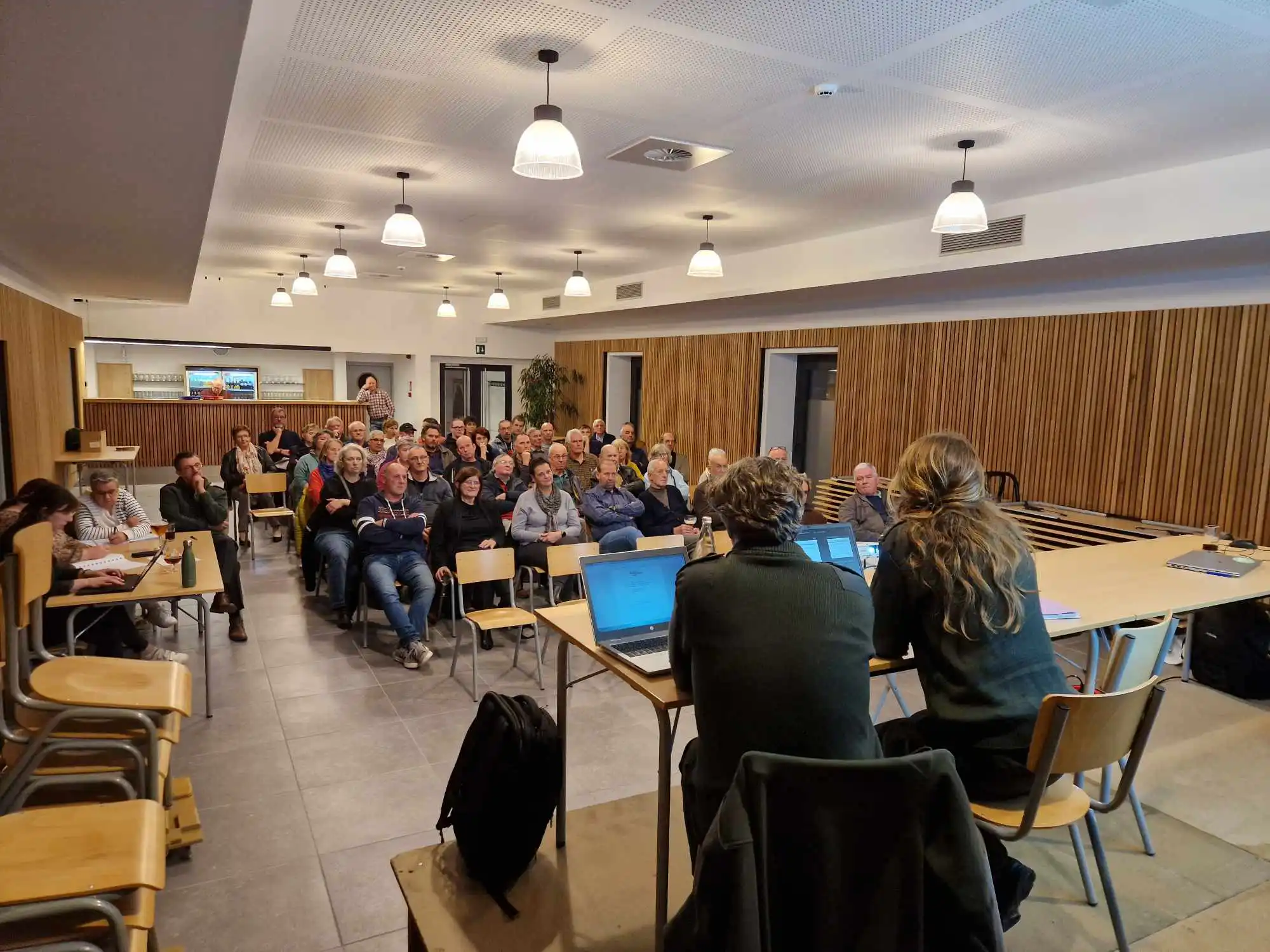 Réunion de la CLDR de Daverdisse. Deux intervenants assis à une table avec des ordinateurs portables, face à un public attentif d’une cinquantaine de personnes. Le public assiste à la présentation d’un projet.