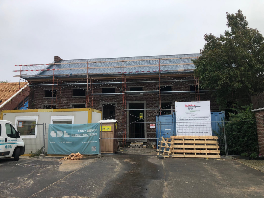 Chantier de rénovation du Mulpas, un bâtiment en briques avec échafaudages sur la façade. Le toit est en cours de réfection, recouvert d’une sous-couche. À l’avant, des containers et panneaux indiquent Constructions. Une zone de stationnement est visible au premier plan
