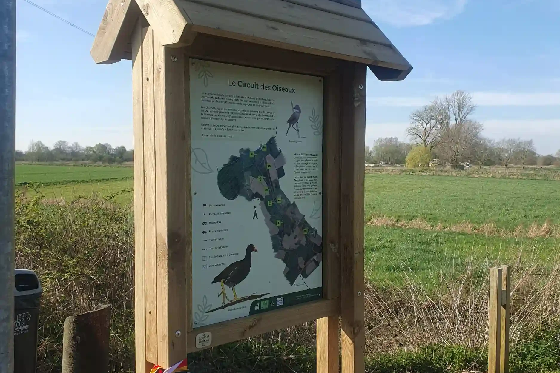 Panneau d'information en bois présentant le “Circuit des Oiseaux”, installé au bord d’un chemin rural entouré de champs et de végétation, sous un ciel bleu légèrement nuageux.