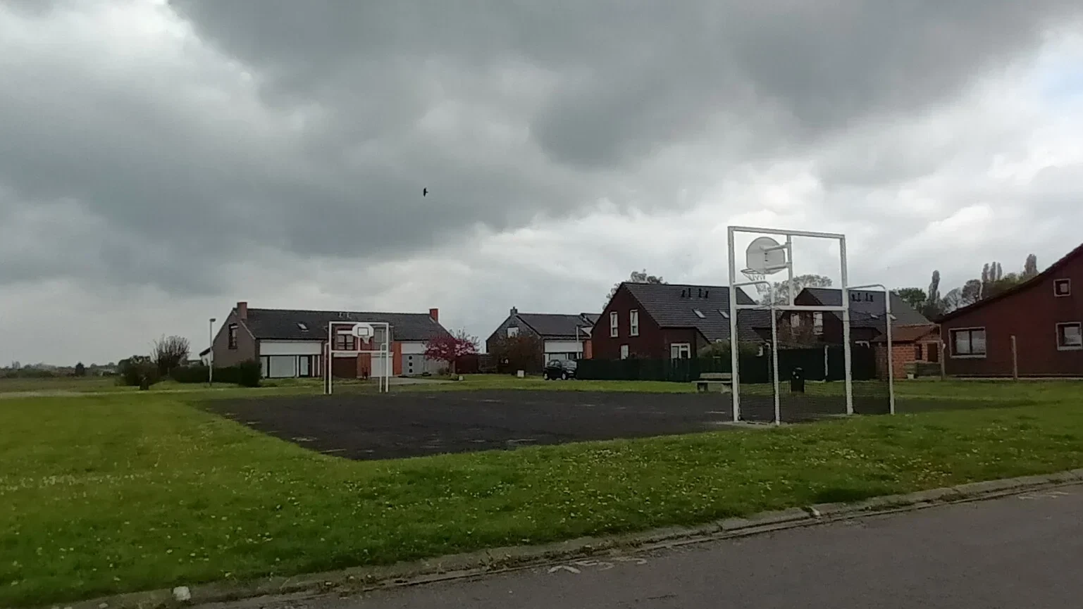 Terrain de basket en plein air au milieu d’un espace vert, avec plusieurs maisons résidentielles en arrière‑plan sous un ciel nuageux.
